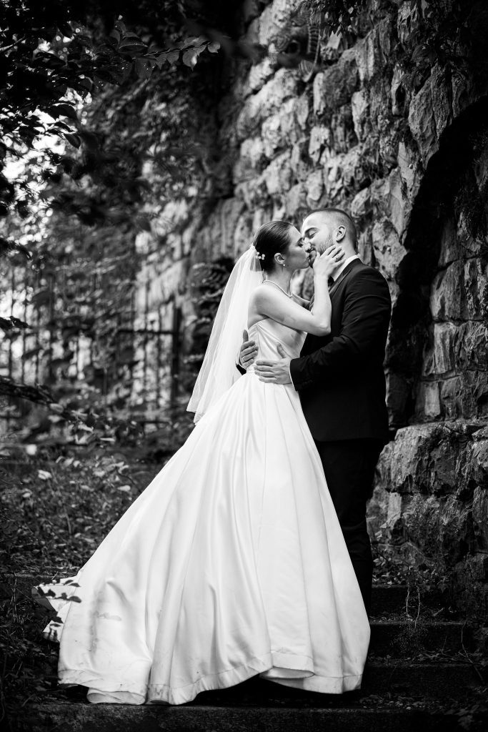 lack and white photo of bride and groom kissing while hugging next to a stone fence