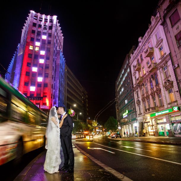 Bride and groom kissing while embracing in downtown Belgrade as a bus rushes by in the background